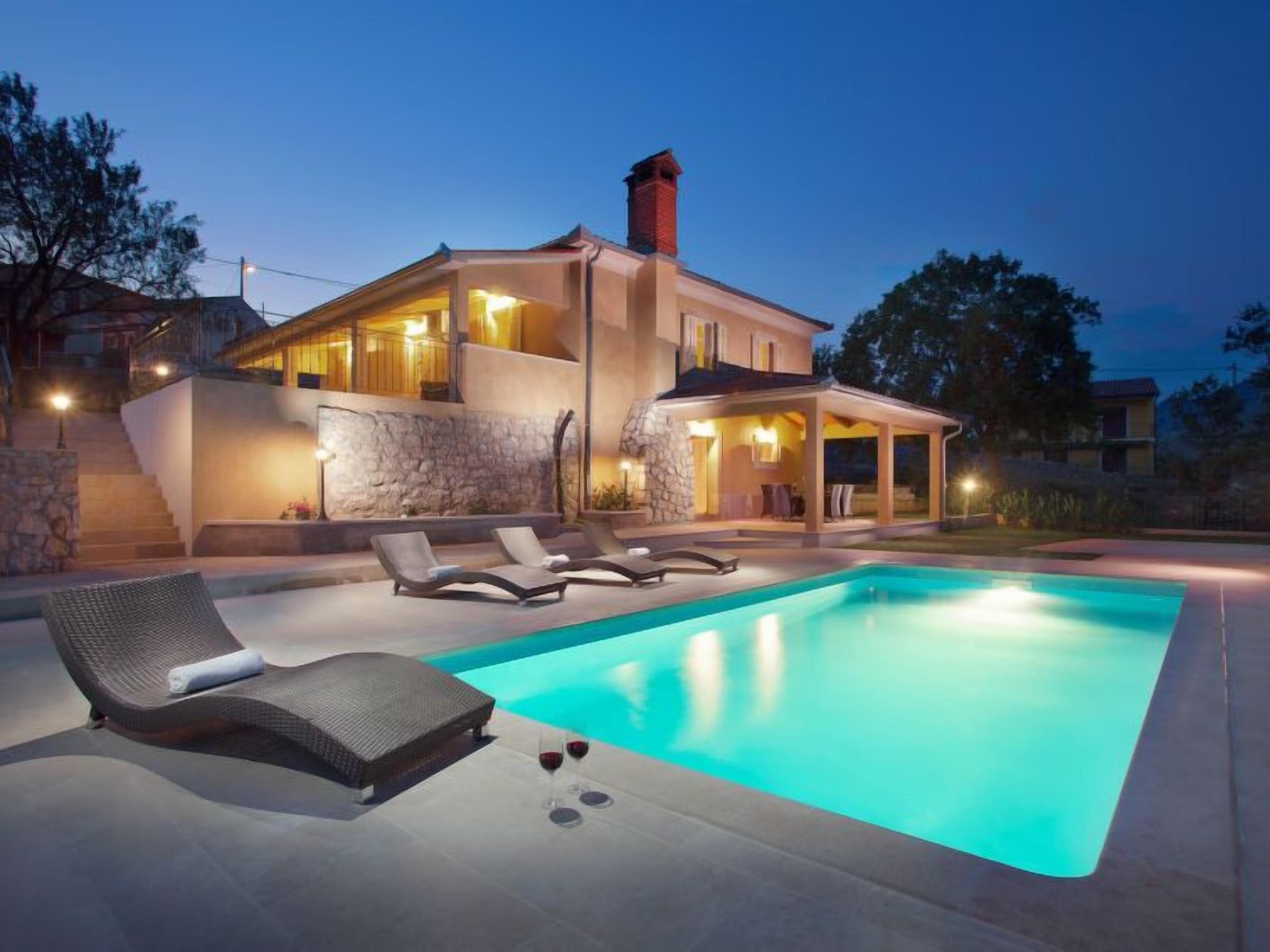 A modern two-story house with a swimming pool, lounge chairs, and a stone wall.