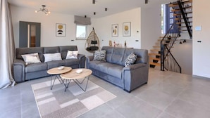 Un salon moderne avec un ensemble de canapés gris, deux petites tables rondes et un fauteuil suspendu. Un escalier avec une rampe noire mène à un niveau supérieur.