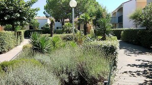 Un camino bordeado por setos cuidados y diversas plantas, que conduce a un edificio con fachada blanca y una lámpara esférica.