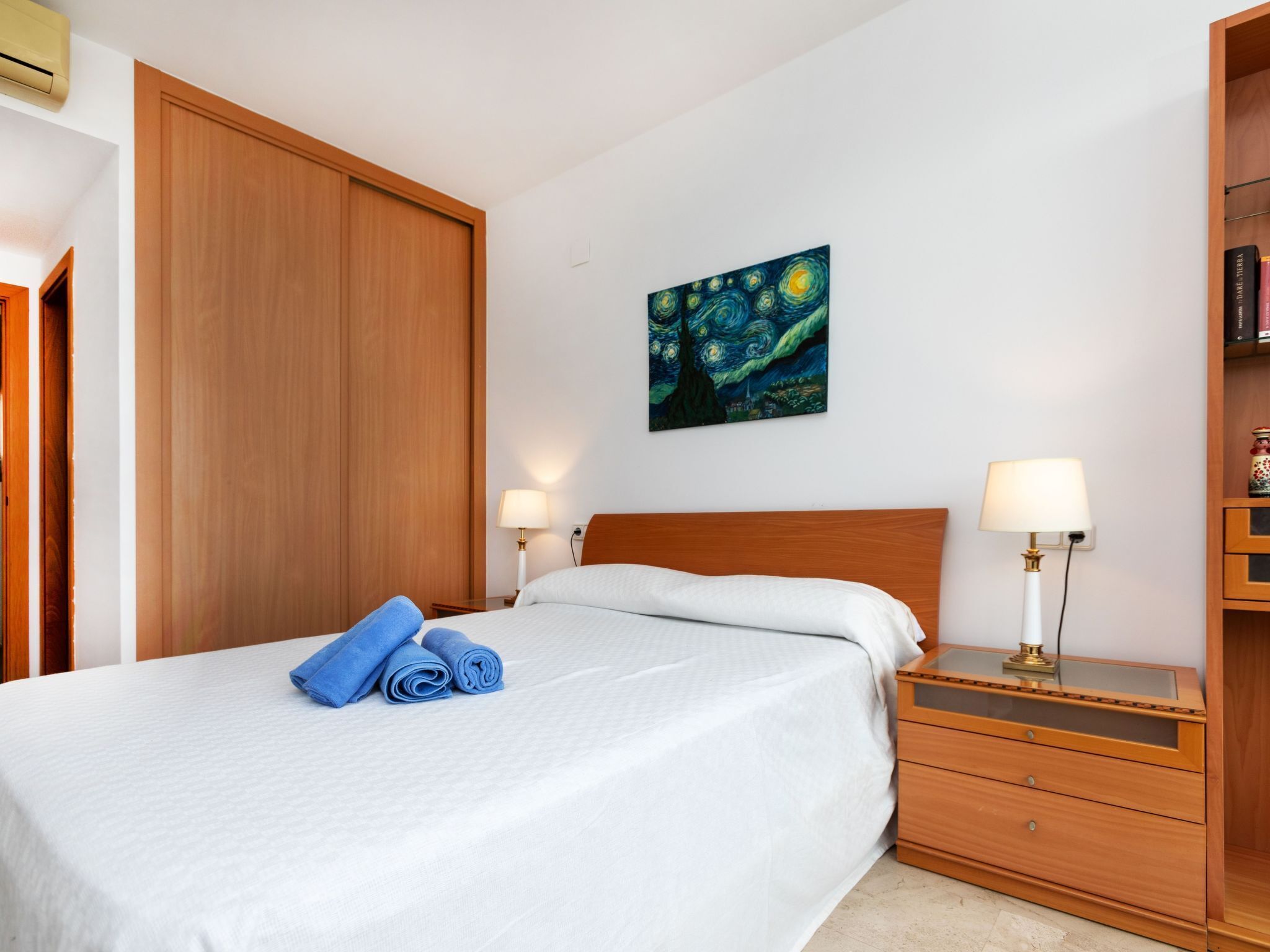 Een net opgemaakt bed met blauwe handdoeken, een houten hoofdbord, een nachtkastje met een lamp en een boekenplank met boeken.