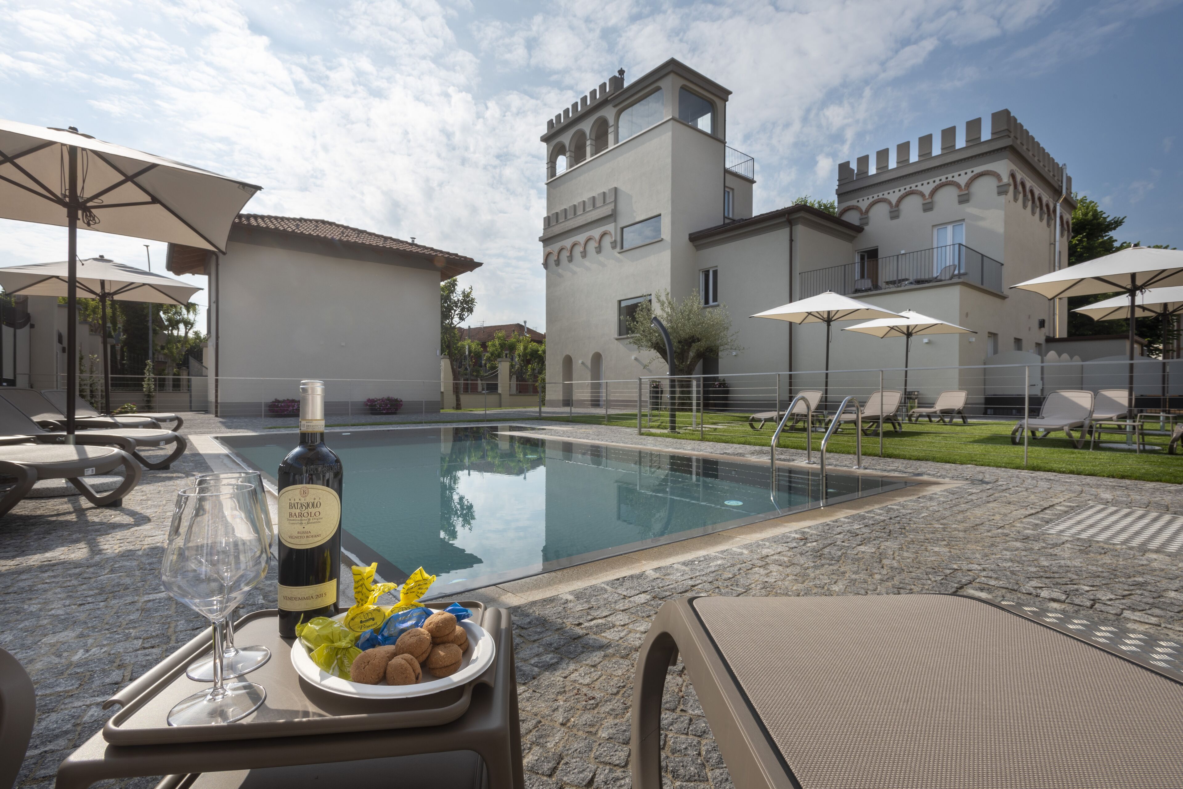 A poolside area with lounge chairs, a table with a bottle of wine and snacks, and a building with a tower-like structure in the background.