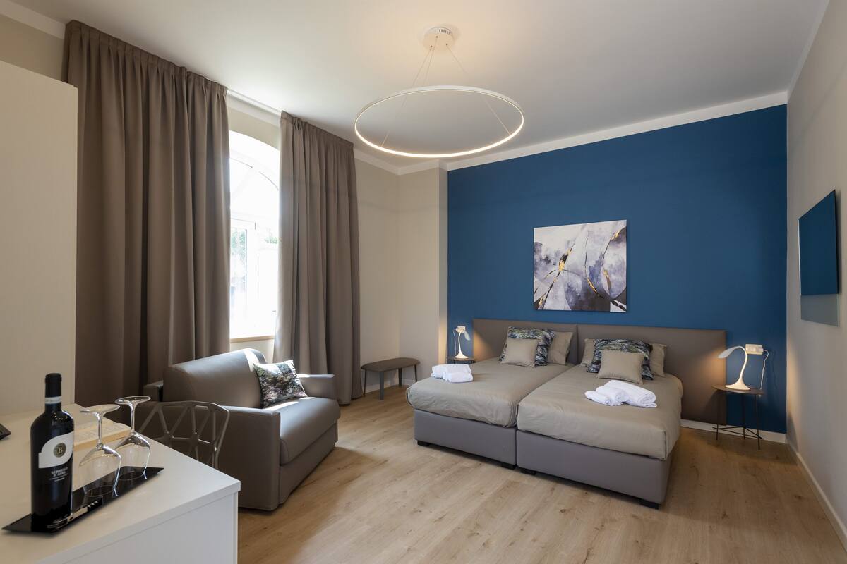 A modern hotel room with a bed, a sofa, a desk with a bottle of wine and glasses, and a painting on the wall.