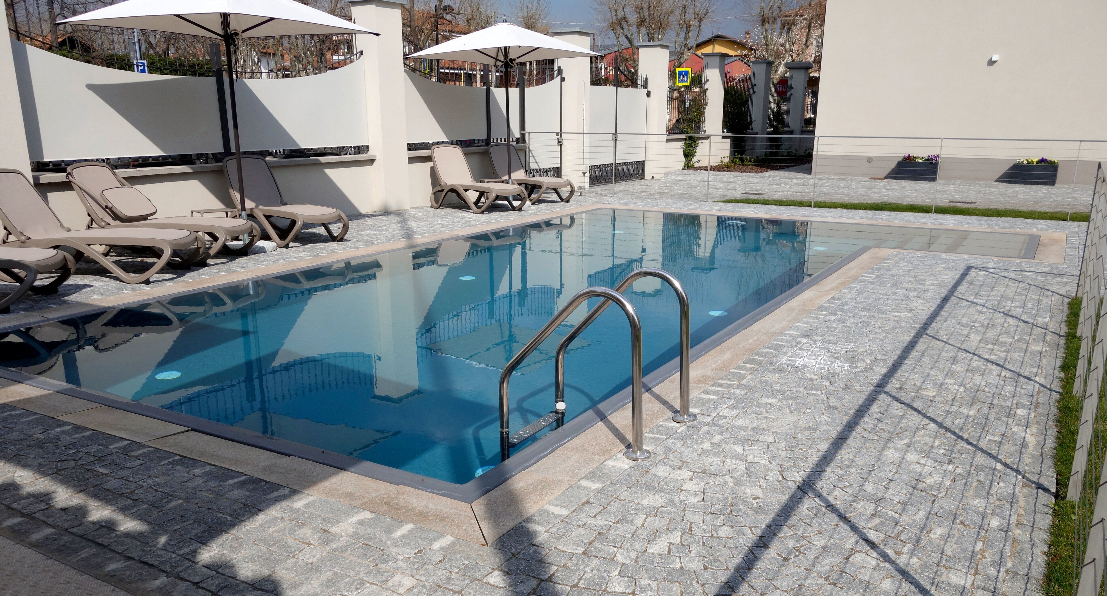 A swimming pool with lounge chairs and umbrellas, surrounded by a paved area and a low wall.
