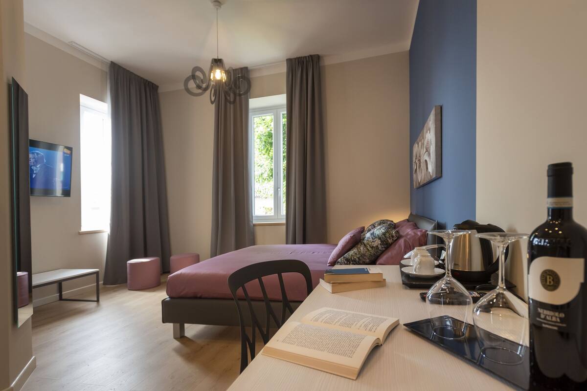 A modern hotel room with a bed, a desk with books and wine glasses, a bench, and a TV.