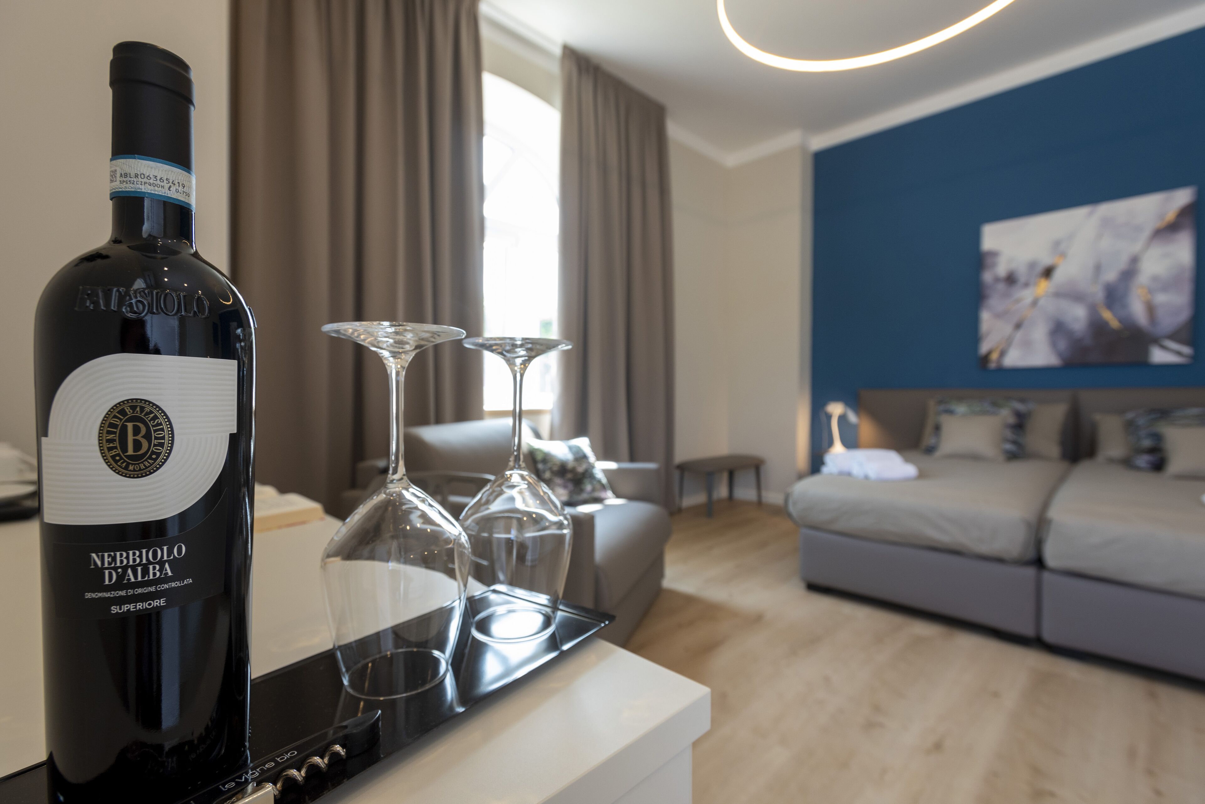 A bottle of Nebbiolo D'Alba wine, two wine glasses, and a modern living room with a sofa and a painting on the wall.