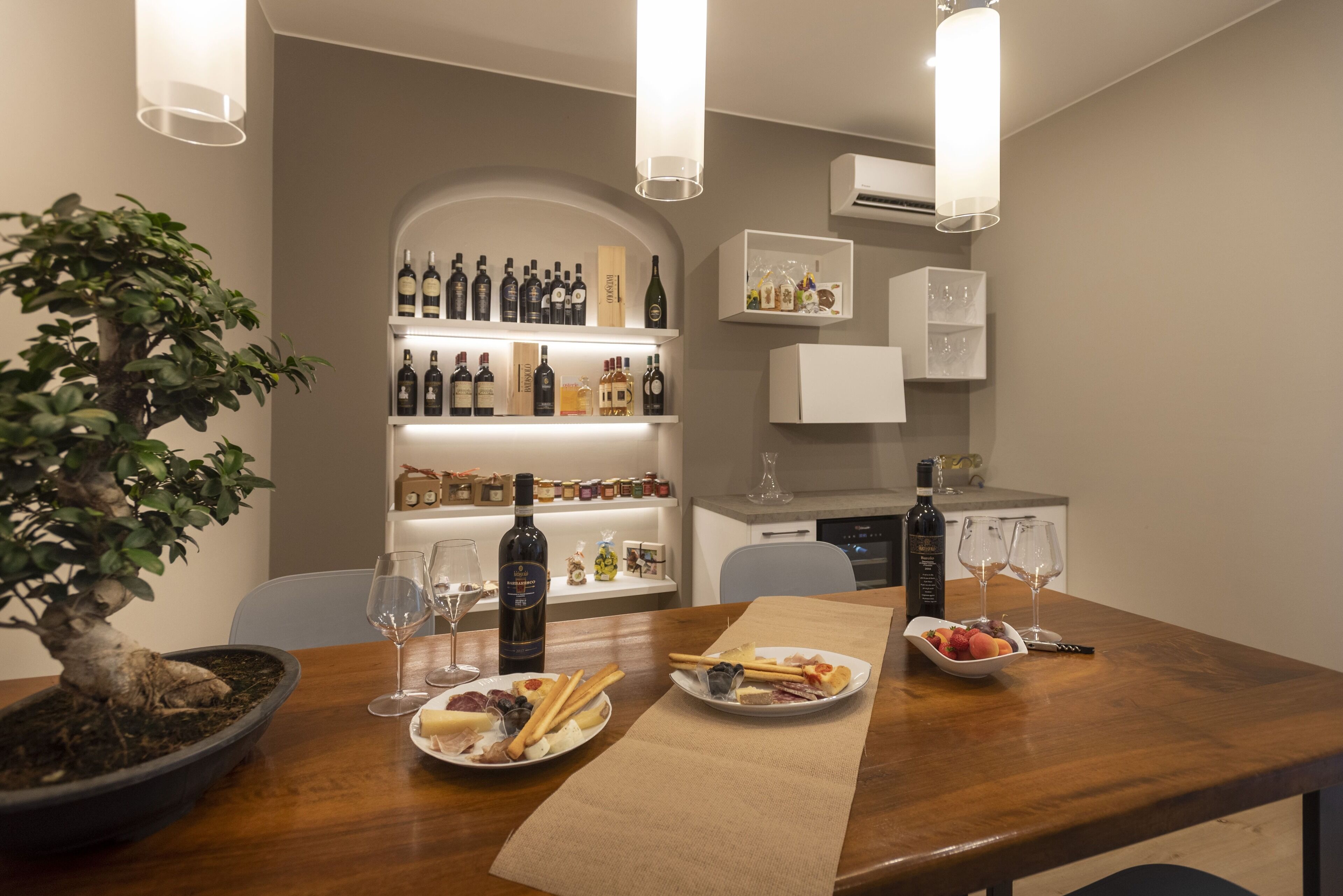 A dining area with a wooden table set for two, featuring plates of food, wine glasses, and a bottle of wine. A bonsai tree is on the left, and shelves with bottles and glasses are in the background.