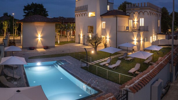 A pool area with lounge chairs and umbrellas, a tower with balconies, and a well-lit building facade. - Villa Bernasconi - camere e ristorante - (Narzole)