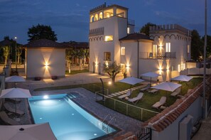 A pool area with lounge chairs and umbrellas, a tower with balconies, and a well-lit building facade. - Villa Bernasconi - camere e ristorante - (Narzole)