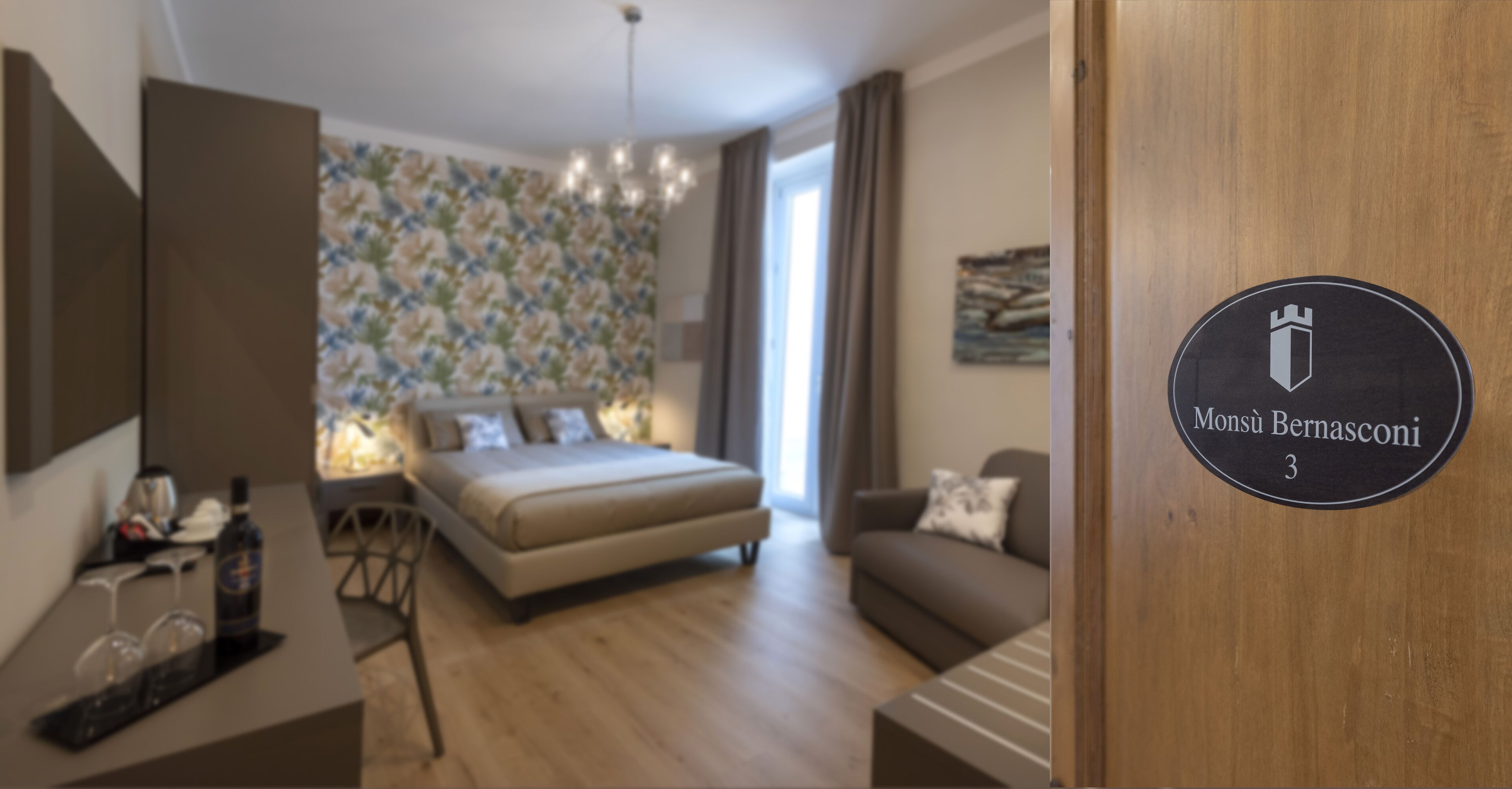 A modern bedroom with a bed, a sofa, a desk, and a chandelier. There is a sign on the door that reads "Monsù Bernasconi 3".