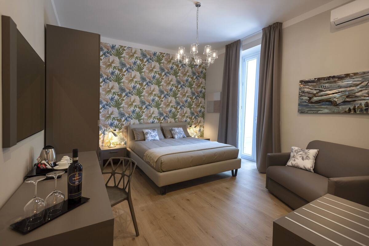 A modern bedroom with a bed, a sofa, a desk with a bottle of wine and glasses, and a wall with a floral pattern.