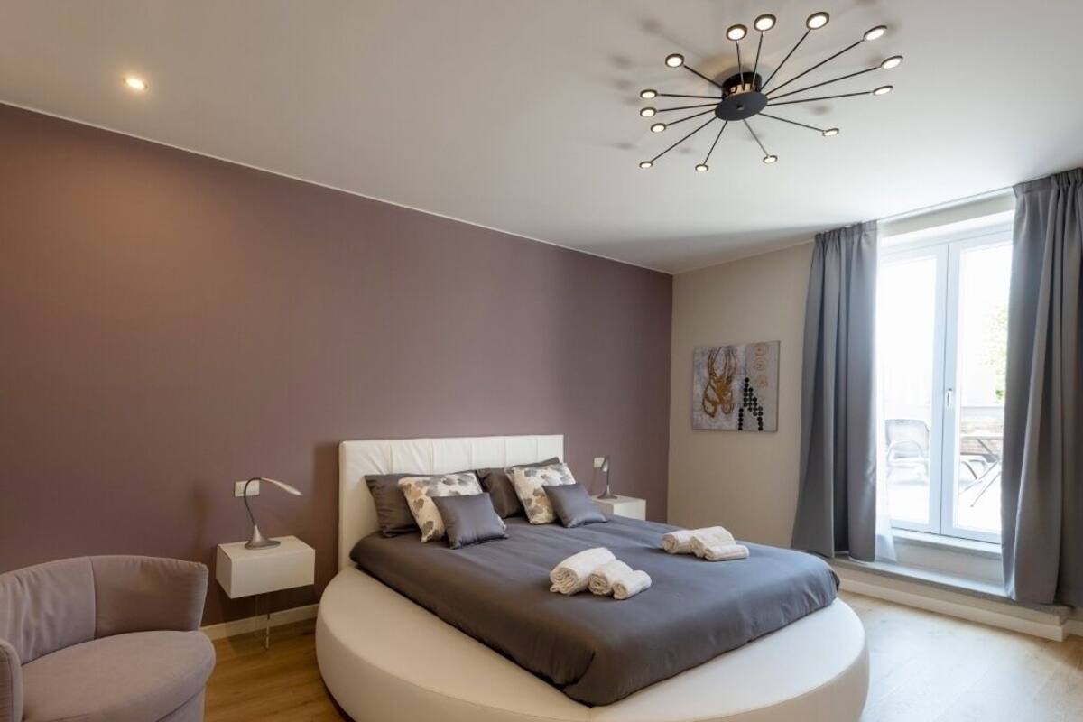 A modern bedroom with a curved bed, a bedside lamp, and a view of the outdoors through a large window.