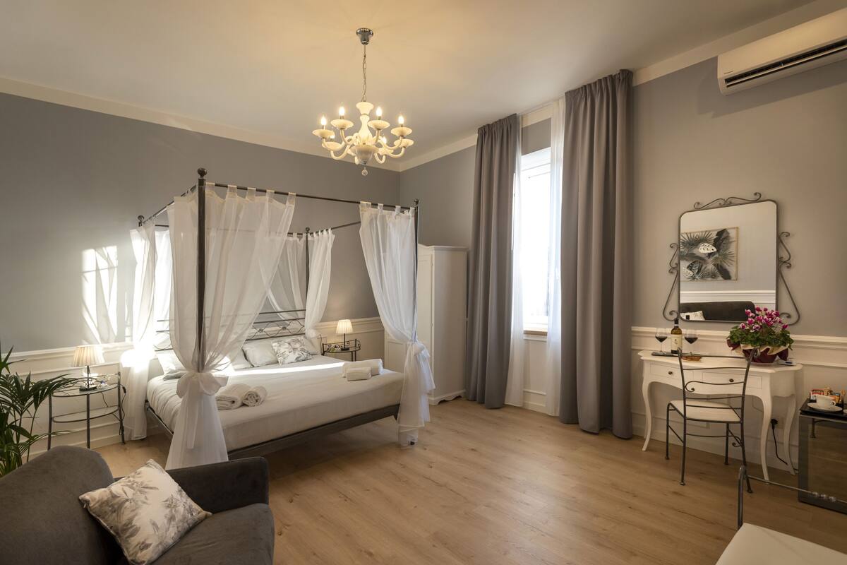 A bedroom with a four-poster bed, a desk with a chair, a small table with a lamp, and a potted plant.