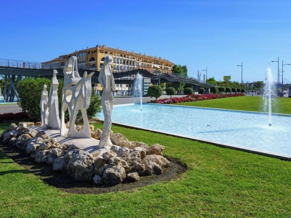A swimming pool with a fountain, surrounded by rocks and grass, with a building in the background.