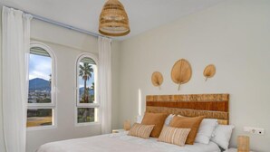 A bedroom with a large bed, two windows with views of palm trees and mountains, a bedside table with a lamp, and a wall-mounted shelf with decorative items.