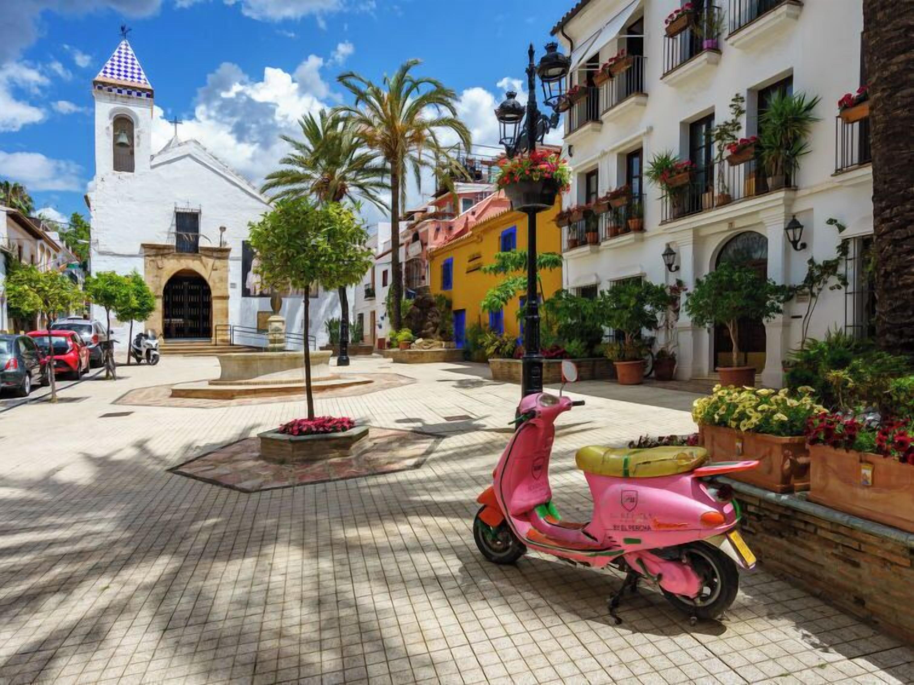A street with a white church, a pink scooter, and buildings with balconies and potted plants.