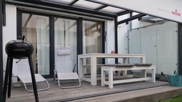 A patio area with a white table and chairs, a black stool, and a glass door leading to a room. - Ferienhaus Breisach by Interhome (Breisach)