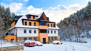 A modern multi-story building with a mix of wooden and white exterior walls, surrounded by snow-covered trees and a clear sky. - Serene Glow Nook & Terrace by Interhome (Pec pod Snezkou)