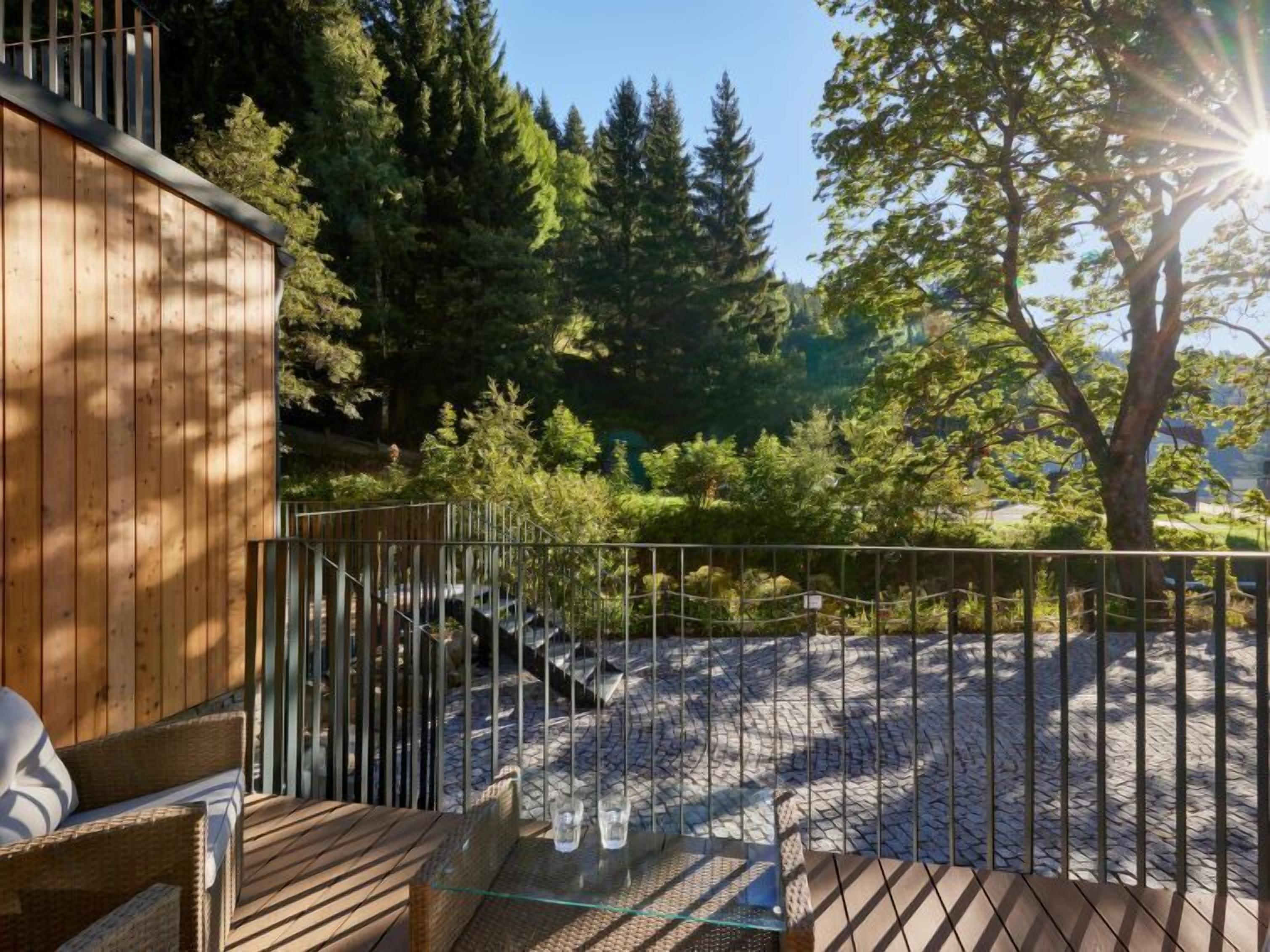 A wooden deck with a glass table and wicker chairs, surrounded by a metal fence and a view of trees and a clear sky.