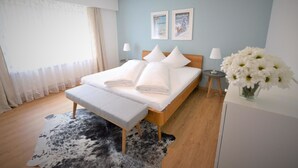 Un dormitorio moderno con una cama de madera, ropa de cama blanca, una pequeña mesita de noche, una mesita auxiliar, un florero con flores y una alfombra de cuero de vaca.