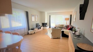 Amplia sala de estar con piso de madera, sofá, televisor y mesa de comedor con sillas.