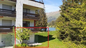 Un edificio moderno de dos pisos con un balcón, un pequeño espacio al aire libre para sentarse y vistas a un lago y montañas.