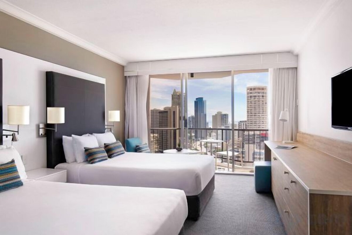 A hotel room with two beds, a desk, a TV, and a balcony with a city view.