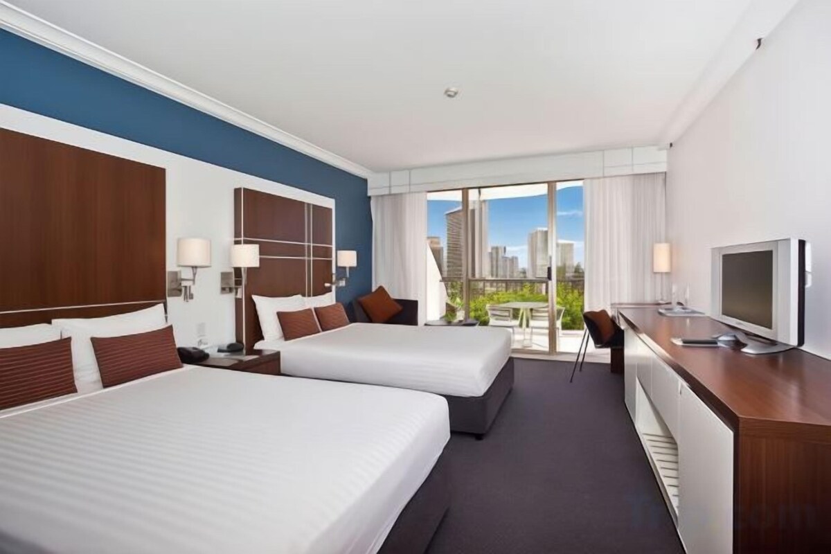A hotel room with two beds, a desk, a TV, and a balcony with a city view.