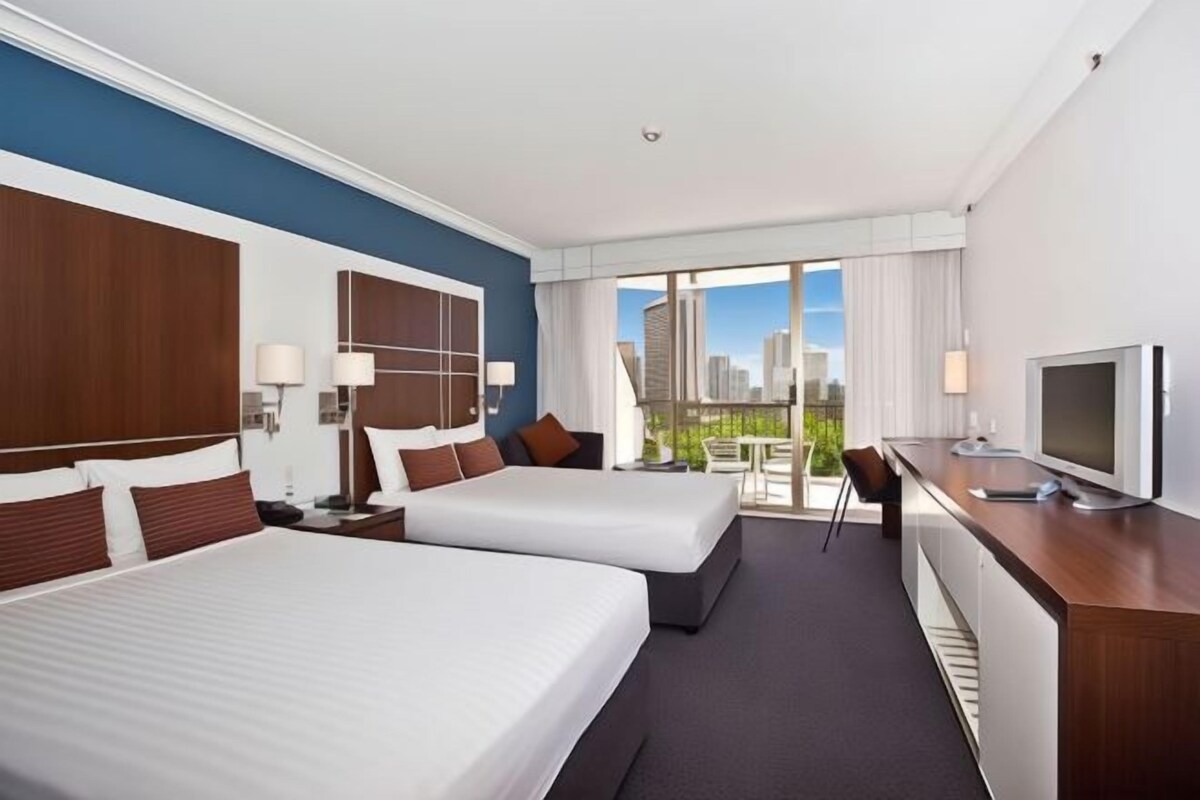 A hotel room with two beds, a desk with a TV, and a balcony with a city view.