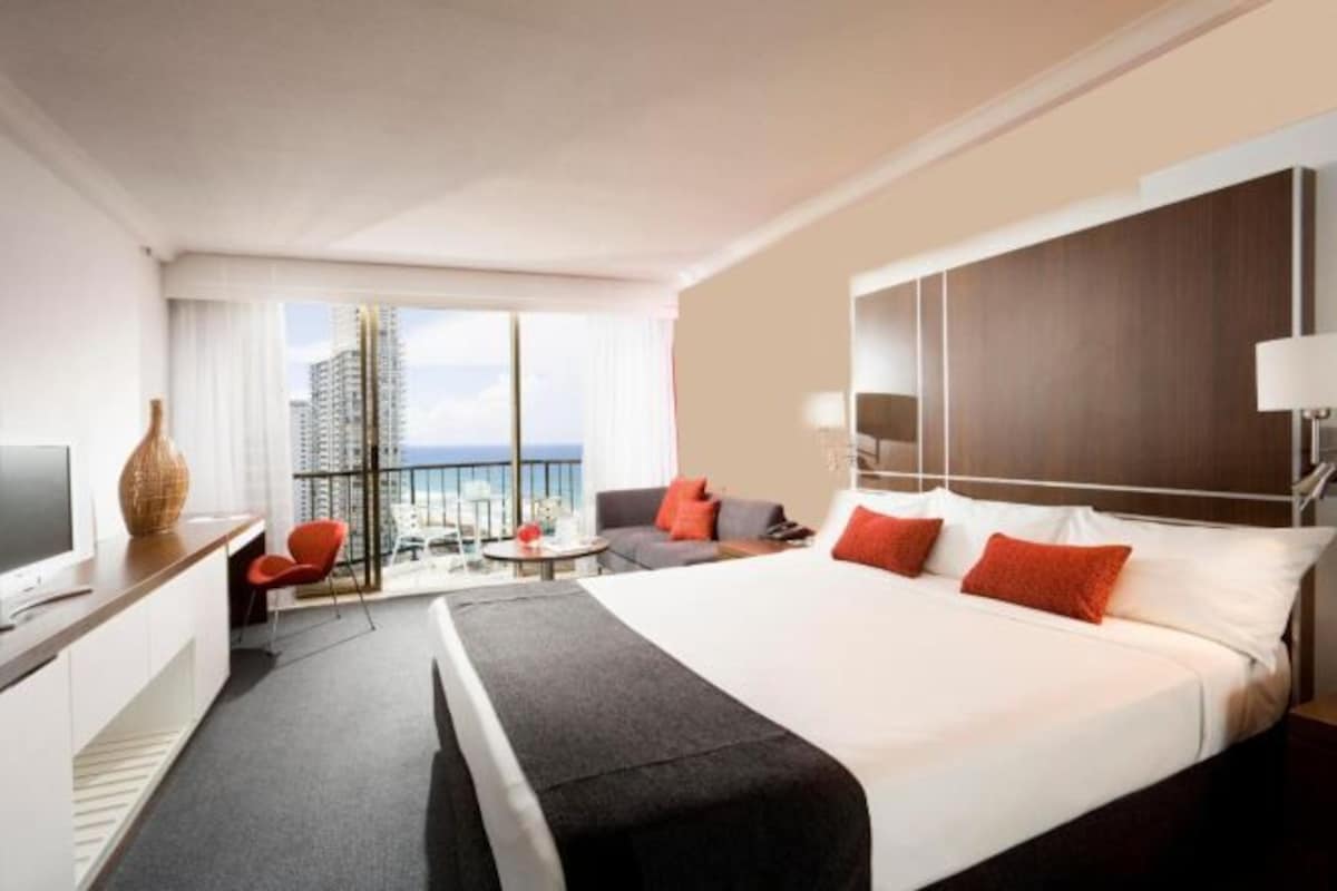 Une chambre d’hôtel dotée d’un grand lit, d’un bureau avec une télévision, d’un coin salon offrant une vue sur la ville et d’un balcon.