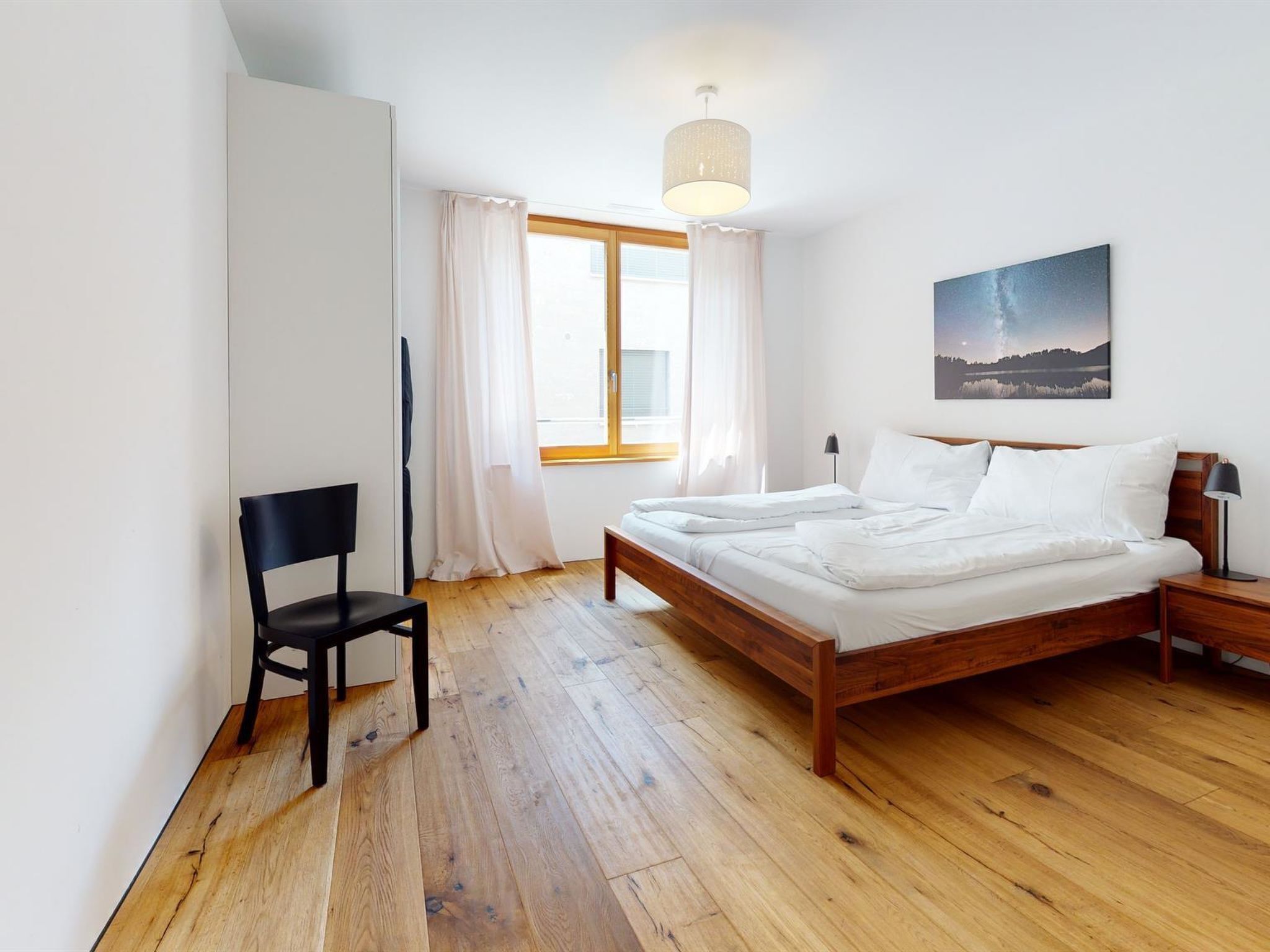 Une chambre à coucher avec un lit en bois, une chaise, une table de chevet, une lampe et une fenêtre avec des rideaux.