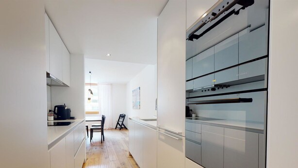 Una cocina moderna con armarios blancos, piso de madera y un rincón comedor con una silla y una mesa.