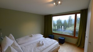 A bedroom with a large bed, two pillows, a bedside table, a window with curtains, and a view of trees and a fence. - Uehlinger Züger by Interhome (Valbella)