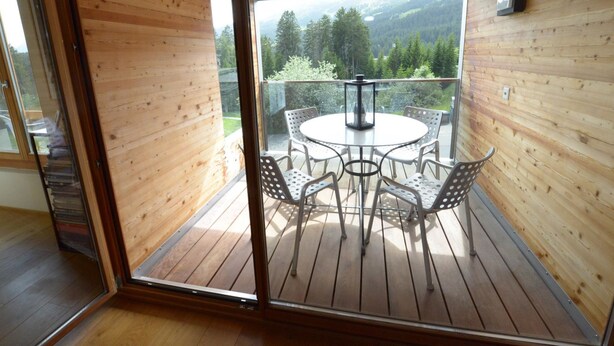 A wooden interior with a balcony featuring a round table and chairs, a lantern, and a view of a forested area. - Uehlinger Züger by Interhome (Valbella)