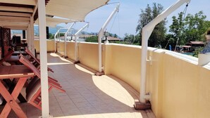 An outdoor terrace with a row of wooden tables and chairs, a yellow wall, and a view of the surrounding landscape. - Neptuna Kata Hotel (Karon)