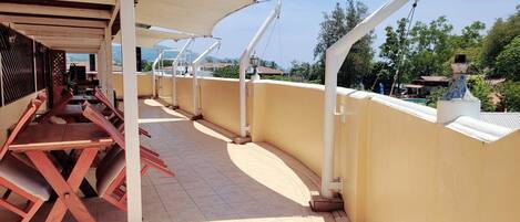 An outdoor terrace with a row of wooden tables and chairs, a yellow wall, and a view of the surrounding landscape.