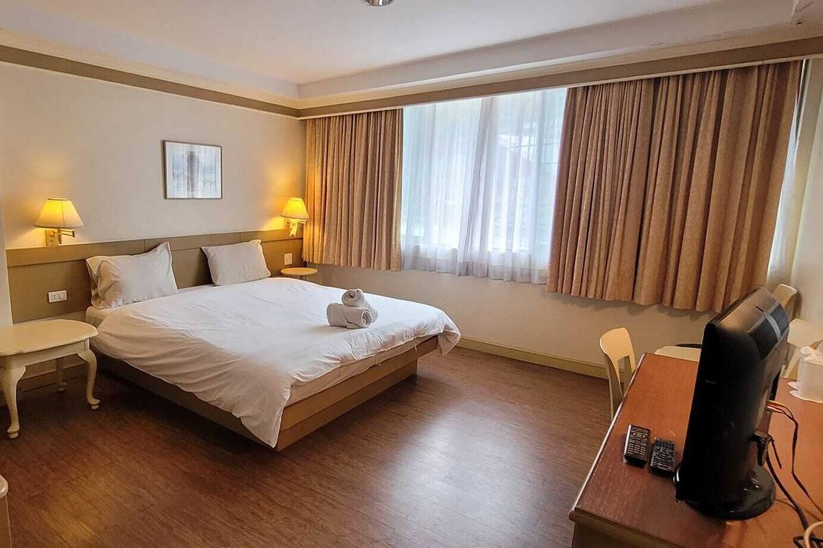A hotel room with a bed, bedside tables, a desk with a computer, and a window with curtains.