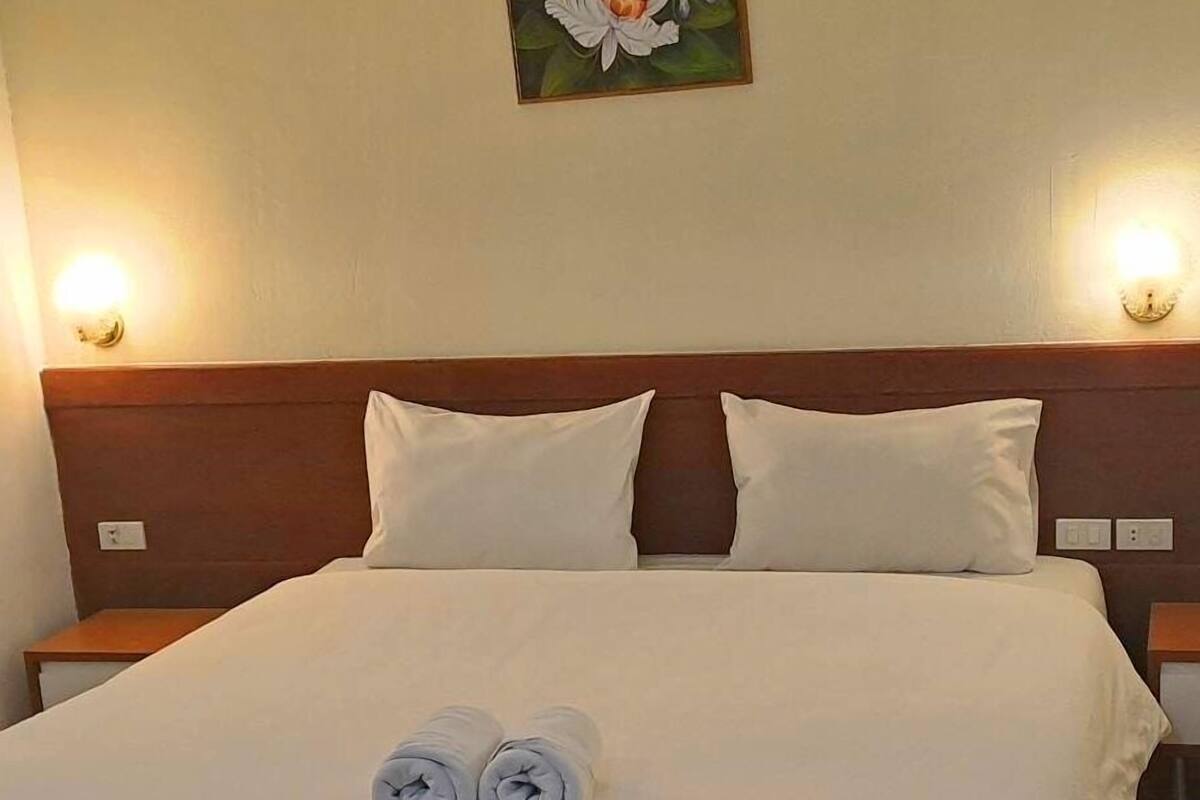 A neatly made bed with white linens and two rolled towels, a wooden headboard, and two bedside lamps.