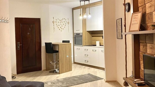 Una sala de estar moderna con un sofá oscuro, una mesa de centro de madera y un mueble de televisión con un televisor de pantalla plana. Hay una zona de cocina con armarios de madera y una mesa de comedor.