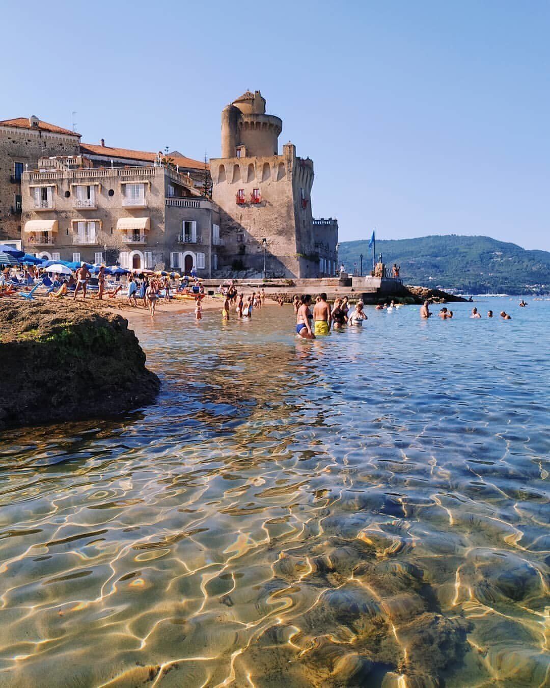 A coastal town with a prominent castle-like building, clear shallow waters, and beachgoers.