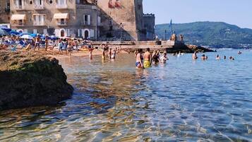 A coastal town with a prominent castle-like building, clear shallow waters, and beachgoers.