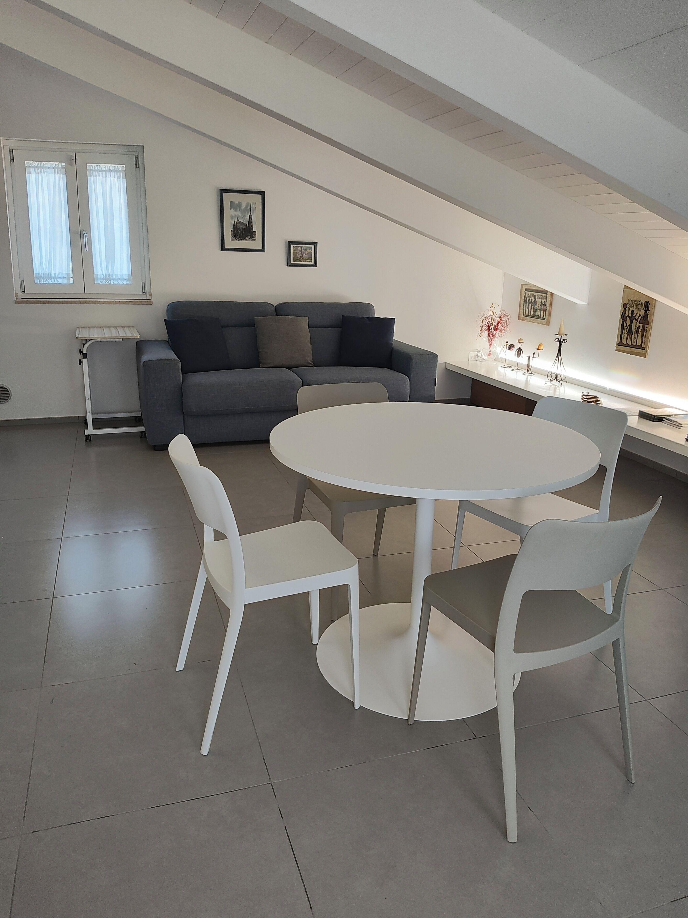 Een moderne woonkamer met een grijze bank, een ronde witte tafel met vier stoelen, en een raam met gordijnen.