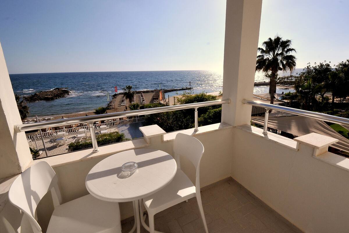 A balcony with a table and chairs, overlooking a beach with a palm tree and a view of the ocean.