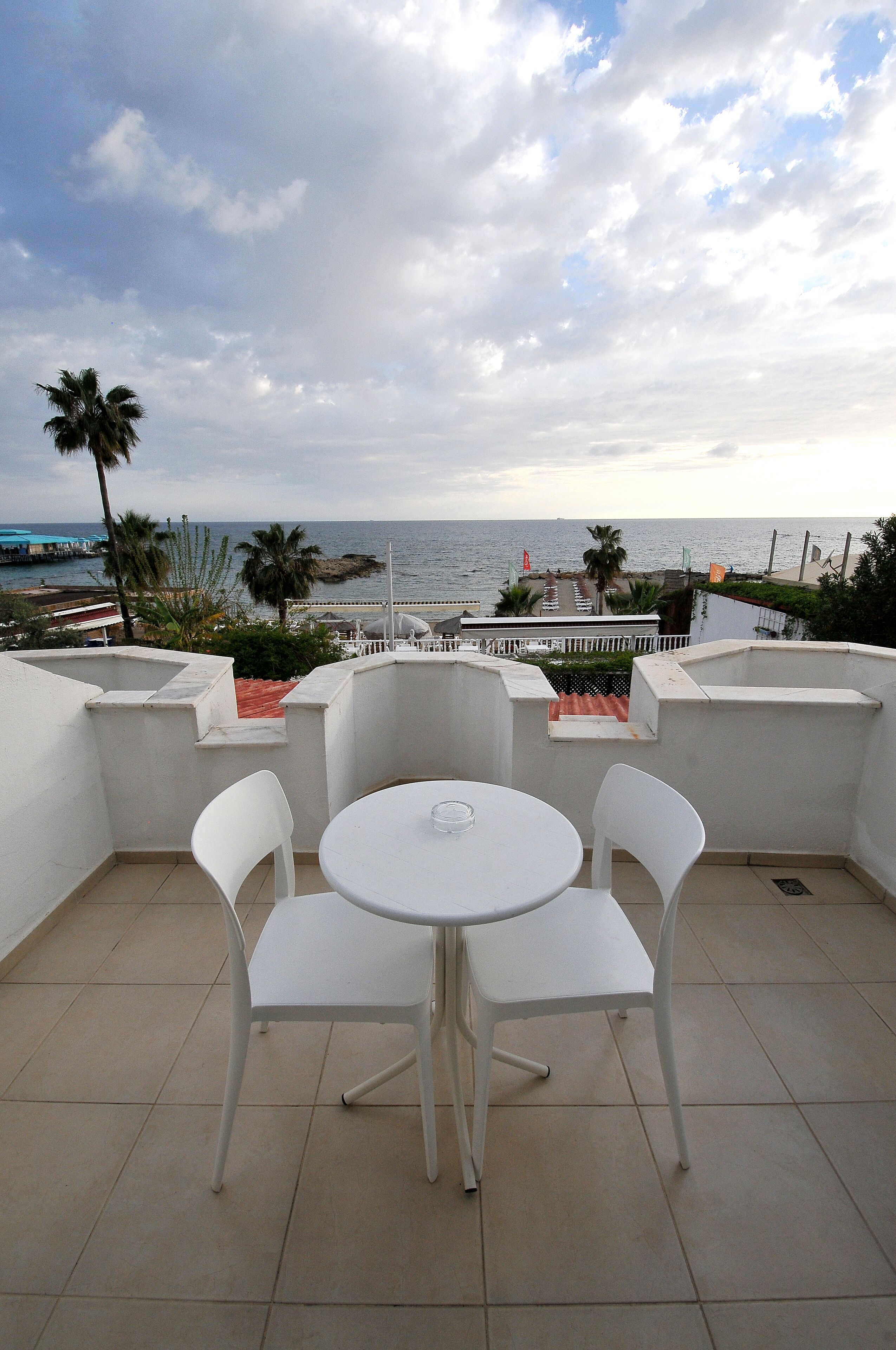 A terrace with a white table and chairs, overlooking a coastal area with palm trees and a view of the sea.