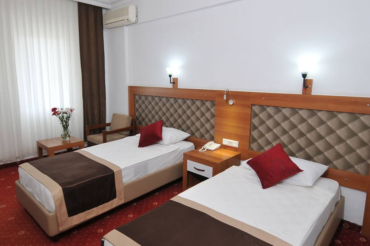 A hotel room with two beds, a bedside table with a lamp, a chair, and a small table with a vase of flowers.