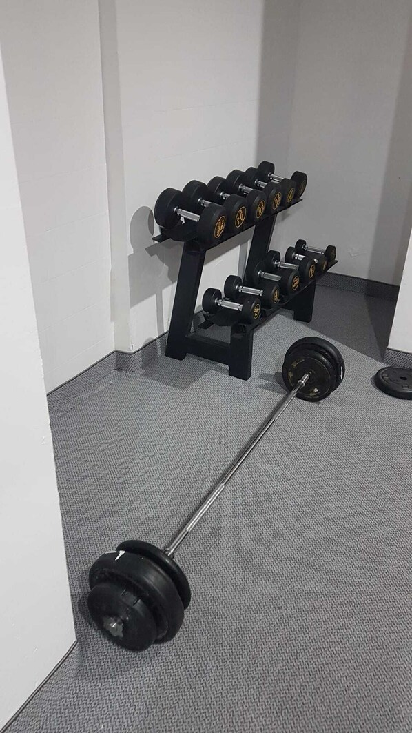 Een sportschool met een halter en gewichtsschijven op de vloer, een dumbbellrek en een witte muur.
