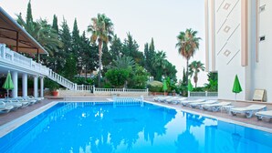 Une piscine entourée de chaises longues et de parasols, bordée de palmiers et d’un bâtiment à la façade blanche.