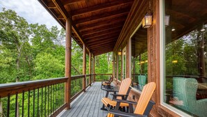 Ett trädäck med Adirondack-stolar och ett glasräcke med utsikt över ett skogsområde.