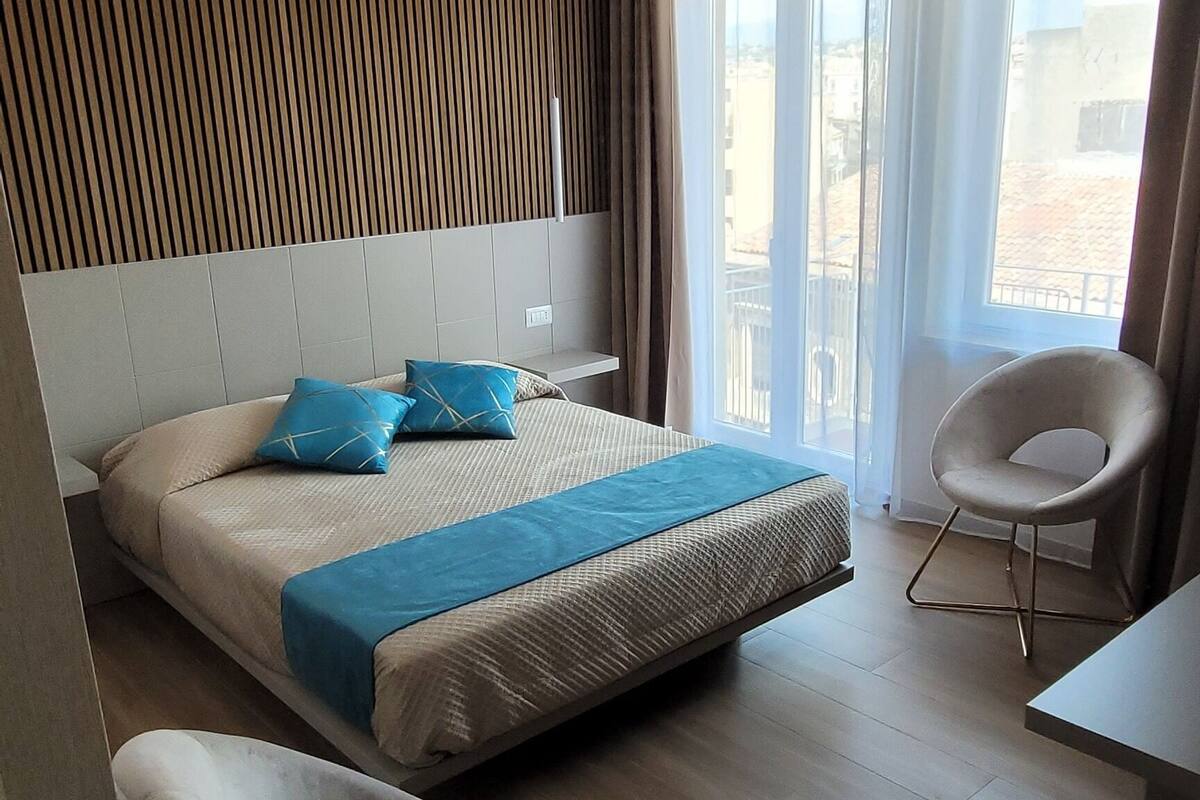 A modern hotel room with a bed, two chairs, a desk, and a view of the outdoors.