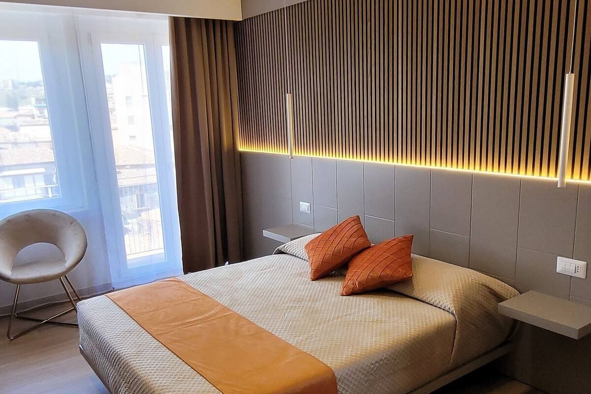 A modern hotel room with a large bed, a chair, and a view through the window.