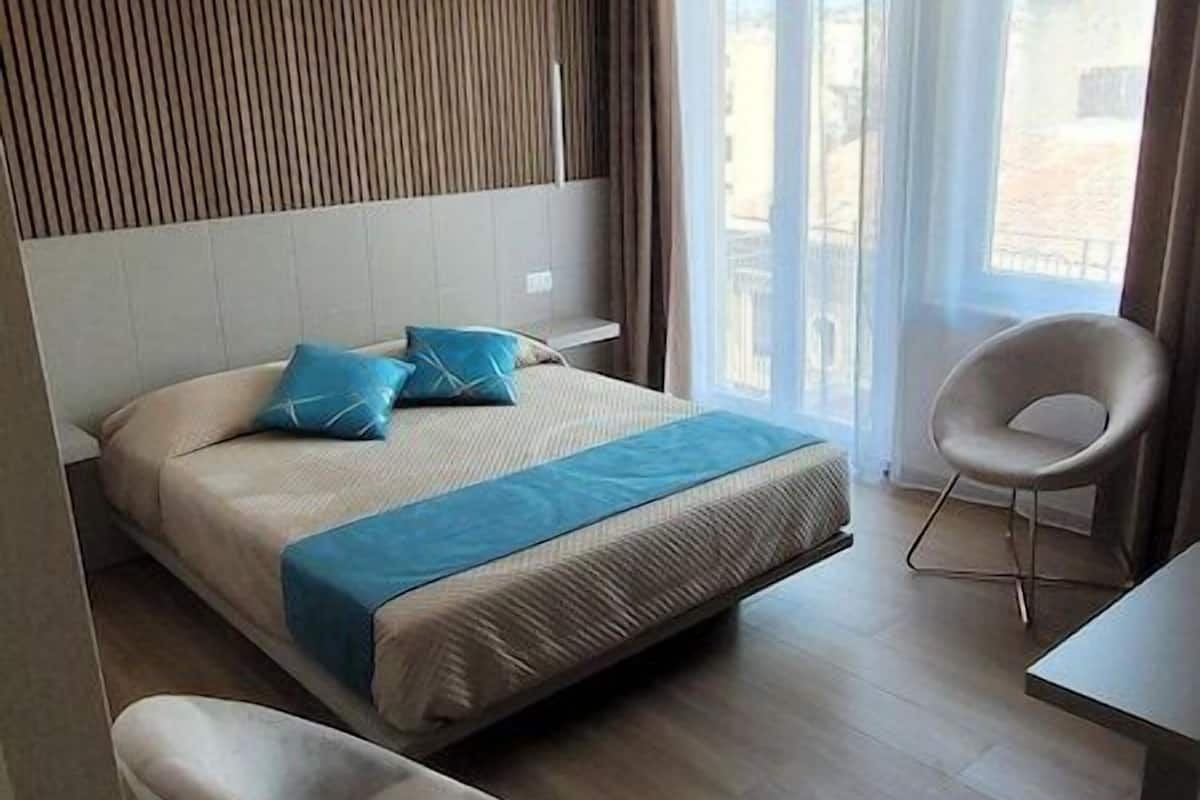 A modern hotel room with a bed, two chairs, a desk, and a large window.